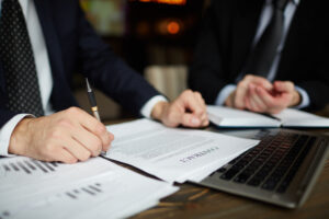 Dois profissionais analisam e assinam um contrato sobre uma mesa, com documentos e um notebook ao lado.